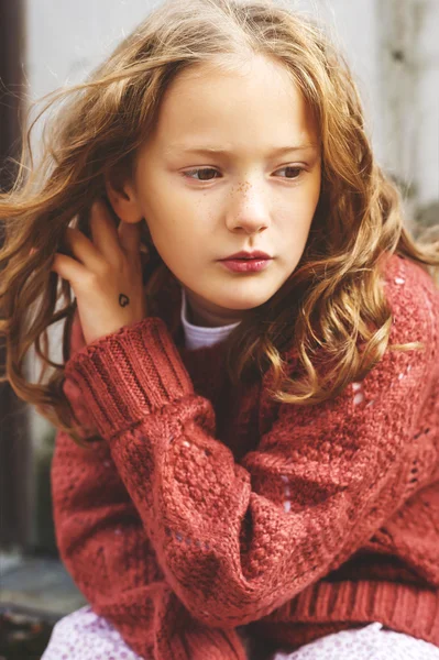 Close up portrait of a cute little girl of 8 years old with curly hair, wearing terracotta pullover, sitting against grey wooden background. Micro contrast toned image — Stock Photo, Image