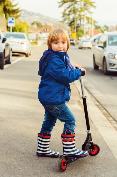 Little boy riding scooter in a city next to big road, wearing blue jacket and stripe rain boots — Stock Photo, Image