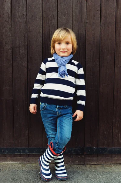 Retrato de moda vertical de menino adorável de 4-5 anos, vestindo listras camisola azul e branca, cachecol, jeans jeans jeans e botas de chuva, de pé contra fundo de madeira marrom escuro — Fotografia de Stock