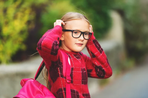 Outdoor portrait of a cute little 9 year old girl, wearing red pullover, glasses and pink backpack — Stock Photo, Image