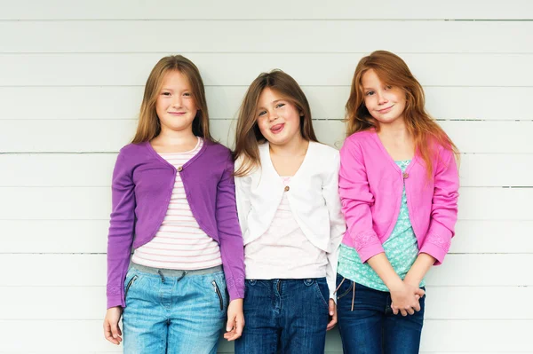 Groep van 3 kleine meisjes permanent buiten tegen witte houten achtergrond, het dragen van denim jeans en kleurrijke jassen — Stockfoto