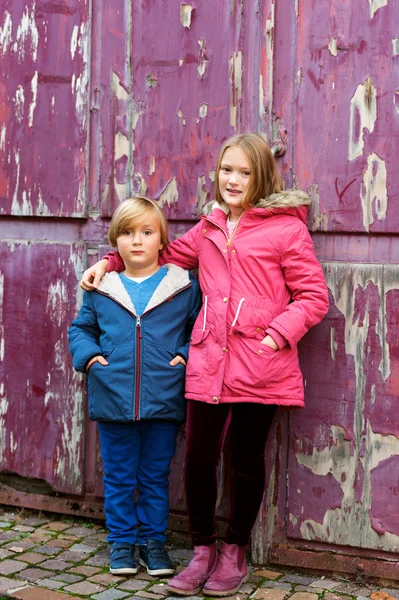 Retrato de duas crianças adoráveis ao ar livre, vestindo casacos quentes em pé ao lado de fundo roxo velho — Fotografia de Stock