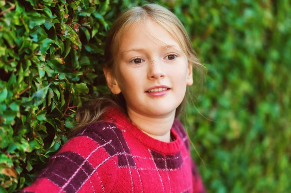 Portrait extérieur en gros plan de mignonne petite fille de 8-9 ans portant un pull rouge vif, debout à côté des buissons verts — Photo