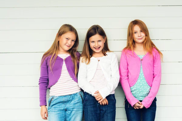 Groep van 3 kleine meisjes permanent buiten tegen witte houten achtergrond, het dragen van denim jeans en kleurrijke jassen — Stockfoto