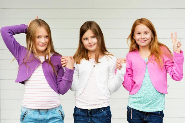 Grupp av 3 små flickor står utomhus mot vit trä bakgrund, klädd i denim jeans och färgglada jackor — Stockfoto
