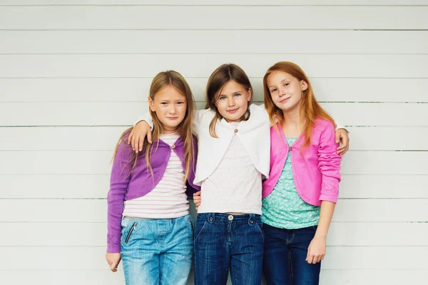 Groep van 3 kleine meisjes permanent buiten tegen witte houten achtergrond, het dragen van denim jeans en kleurrijke jassen — Stockfoto