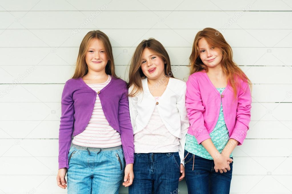 Group of 3 little girls standing outdoors against white wooden background, wearing denim jeans and colorful jackets