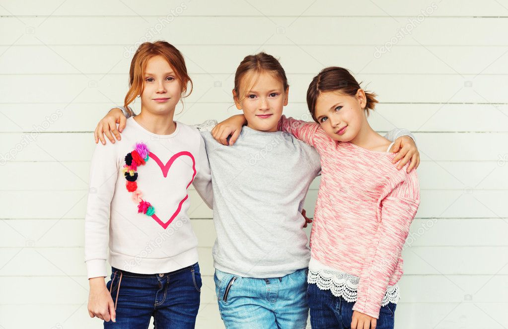 Group of 3 little girls standing outdoors against white wooden background, wearing denim jeans and sweatshirts