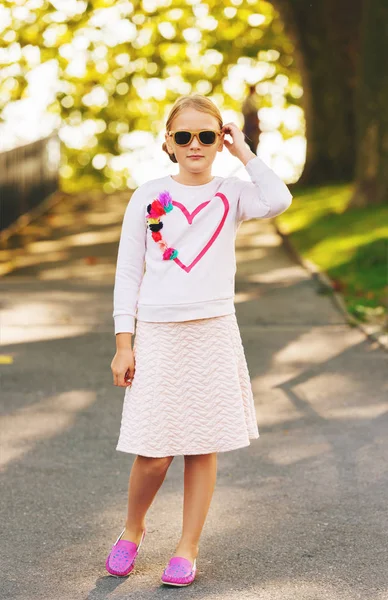 Modeporträt eines niedlichen kleinen Mädchens im Freien, das ein weißes Sweatshirt mit Herz, rosa Rock und Mokassins-Schuhen trägt — Stockfoto