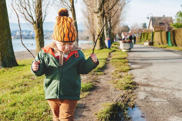 Симпатичный 3-летний мальчик, играющий на улице в солнечный ранний весенний день, в зеленой куртке и оранжевой шляпе — стоковое фото