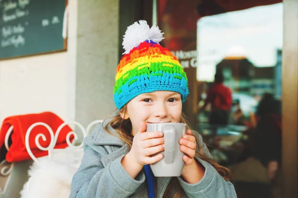 Adorável menina de 6 anos bebendo chocolate quente no café de inverno, segurando copo grande, vestindo casaco cinza e chapéu colorido — Fotografia de Stock