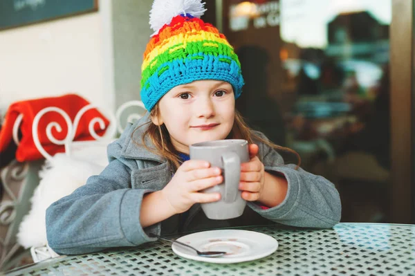Adorável menina de 6 anos bebendo chocolate quente no café de inverno, segurando copo grande, vestindo casaco cinza e chapéu colorido — Fotografia de Stock