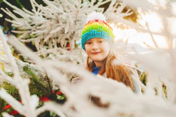 Söt liten flicka klädd i färgglada hatt, stående mellan julgranar vid solnedgången — Stockfoto