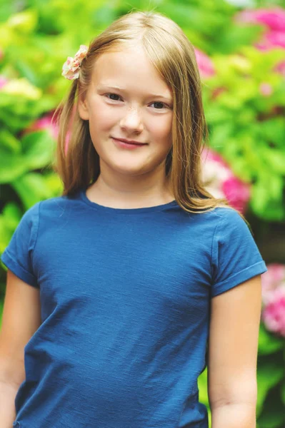 Outdoor vertical portrait of a cute little girl of 9 years old in summer garden, leaning on a fence, wearing blue tee shirt — Stock Photo, Image