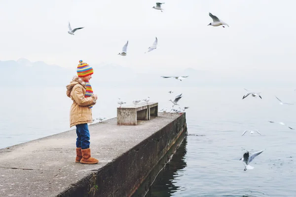 Милая девочка играет на открытом воздухе рядом с Женевским озером в холодную погоду, кормит птиц в зимнее время — стоковое фото