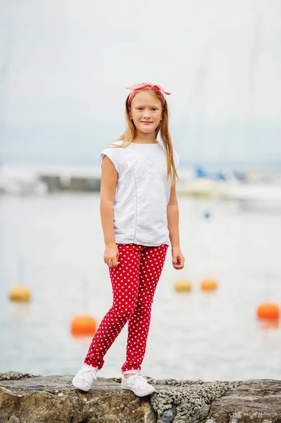 Счастливая 9-летняя девочка, весело проводящая время на свежем воздухе, играющая у озера приятным теплым солнечным вечером, в белой футболке и туфлях, красных брюках-польке и головном уборе. Фото сделано на озере Женева, Швейцария — стоковое фото