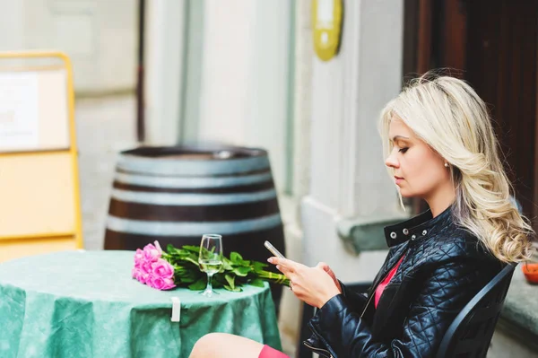 Ritratto all'aperto di bella donna bionda che tiene bouquet di rose rosa, riposando nel caffè, utilizzando lo smartphone — Foto Stock