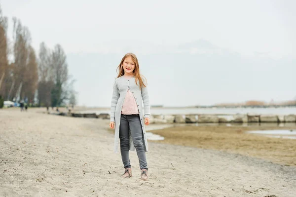Adorable little girl of 8-9 years old playing by the lake, wearing grey trousers and long cardigan — Stock Photo, Image