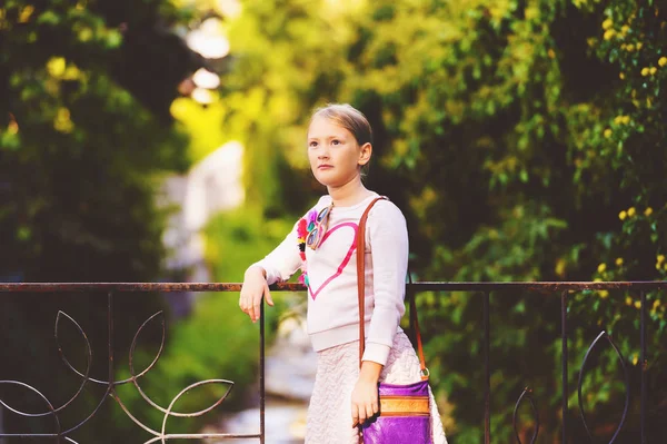 Fashion portrait of a cute little girl outdoors, wearing white sweatshirt with heart and purple bag — Stock Photo, Image