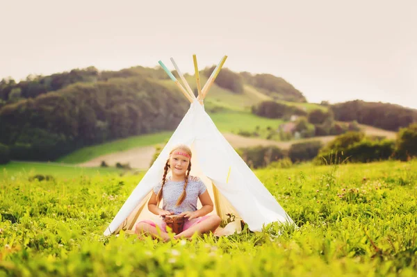 Cute little girl having fun outdoors, playing with tambour, sitting next to teepee — Stock Photo, Image