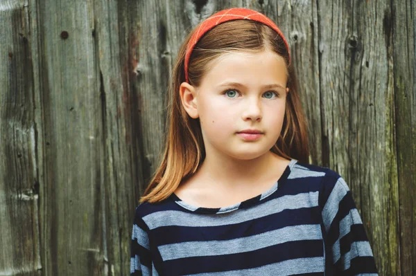 Außenporträt eines niedlichen kleinen 8-9 jährigen Mädchens mit braunen Haaren, das ein gestreiftes Oberteil trägt und vor altem Holzhintergrund steht — Stockfoto