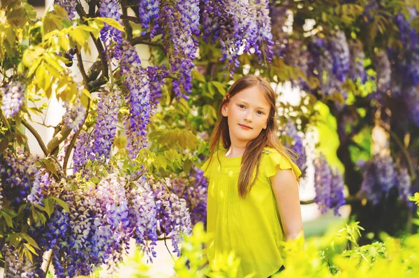 Buiten stijlvolle portret van een schattig klein meisje van 8-9 jaar oud, permanent naast prachtige paarse blauweregen bloemen, groene blouse dragen — Stockfoto