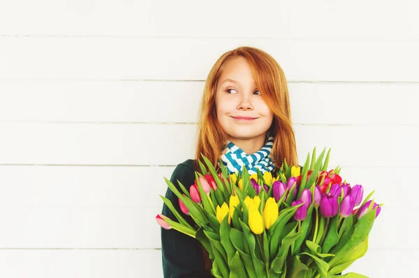 Portrait of redhead girl of 8-9 years old, holding bright bouquet of colorful fresh tulips, standing against white wooden background — Stock Photo, Image