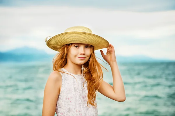 Pretty little girl with long red hair playing by the lake on a very windy day, wearing big hat — Stock Photo, Image
