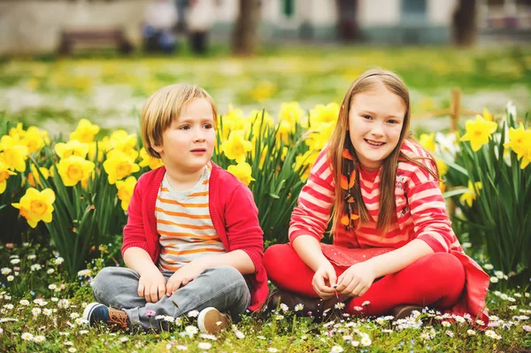 Deux mignons enfants, petit garçon et sa grande sœur, jouant dans le parc entre des fleurs jaunes de jonquilles, portant des vêtements rouge vif — Photo