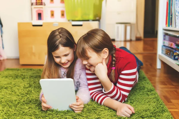 Due bambine carine che giocano sul tablet digitale, sdraiate sul tappeto verde nella stanza dei bambini — Foto Stock