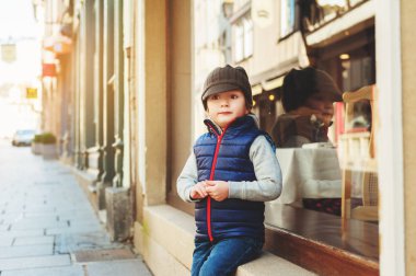 Strasbourg, Fransa sokaklarında oturan çocuk. Mavi yelek, denim jeans, kahverengi şapka giyen çocuk 