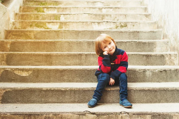 Entzückender kleiner Junge sitzt auf einer Treppe in einer Stadt, trägt roten und blauen Pullover, Jeans und Mokassins Schuhe — Stockfoto