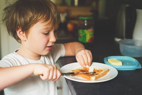 Bonito menino de 6 anos preparando sozinho sua torrada com manteiga — Fotografia de Stock