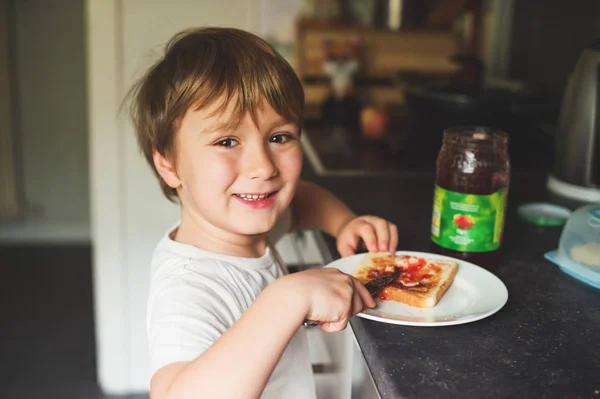 Bonito menino de 6 anos preparando sozinho sua torrada com manteiga e geléia — Fotografia de Stock