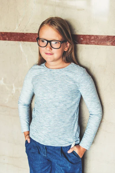 Interior portrait of a cute school girl wearing glasses, stripy shirt, denim jeans, hands in pockets — Stock Photo, Image