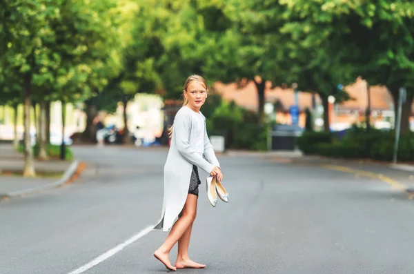 Little barefoot girl walking down the street, wearing long grey cardigan, holding silver bow ballerinas shoes — Stock Photo, Image