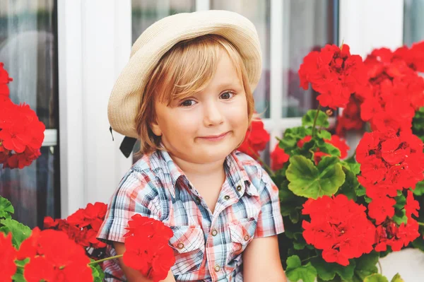 Adorable little boy gardener sitting between bright red geranium in pots — Stock Photo, Image