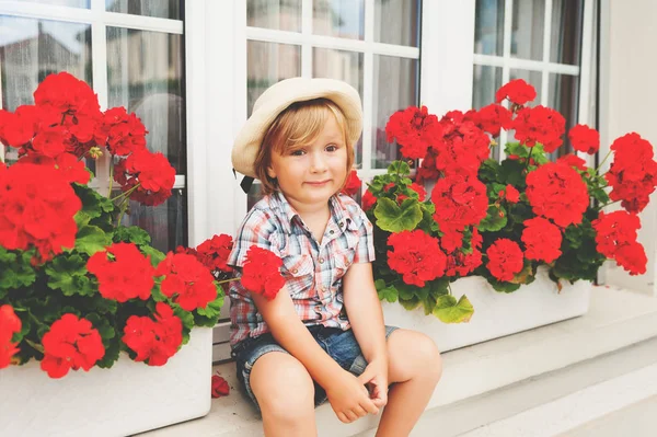 Adorable little boy gardener sitting between bright red geranium in pots — Stock Photo, Image