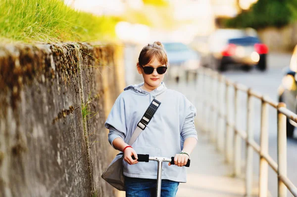 Feliz menina da escola de 9 anos com mochila nas ruas da cidade, andando de scooter, usando óculos de sol — Fotografia de Stock