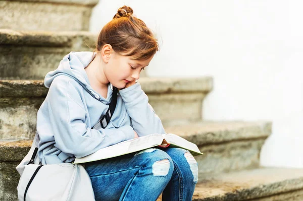 Kid girl reading book outside, sitting on stairs, wearing grey sweatshirt and backpack — Stock Photo, Image