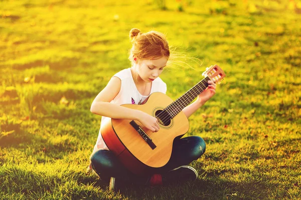 Plenerowej urocza 9-letniej dziewczynki dziecko grać na gitarze na zewnątrz, siedząc na jasny zielony trawnik w ciepły, letni zachód słońca — Zdjęcie stockowe