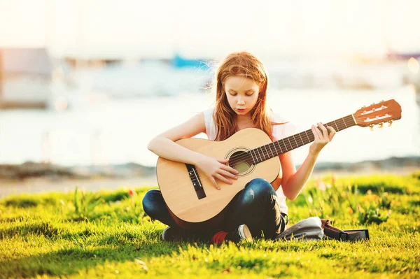 Plenerowej urocza 9-letniej dziewczynki dziecko grać na gitarze na zewnątrz, siedząc na jasny zielony trawnik w ciepły, letni zachód słońca — Zdjęcie stockowe
