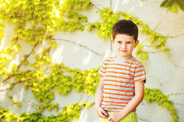 Retrato al aire libre de un lindo niño parado contra la pared de hiedra en un día muy soleado, usando una camiseta de rayas gris y naranja —  Fotos de Stock