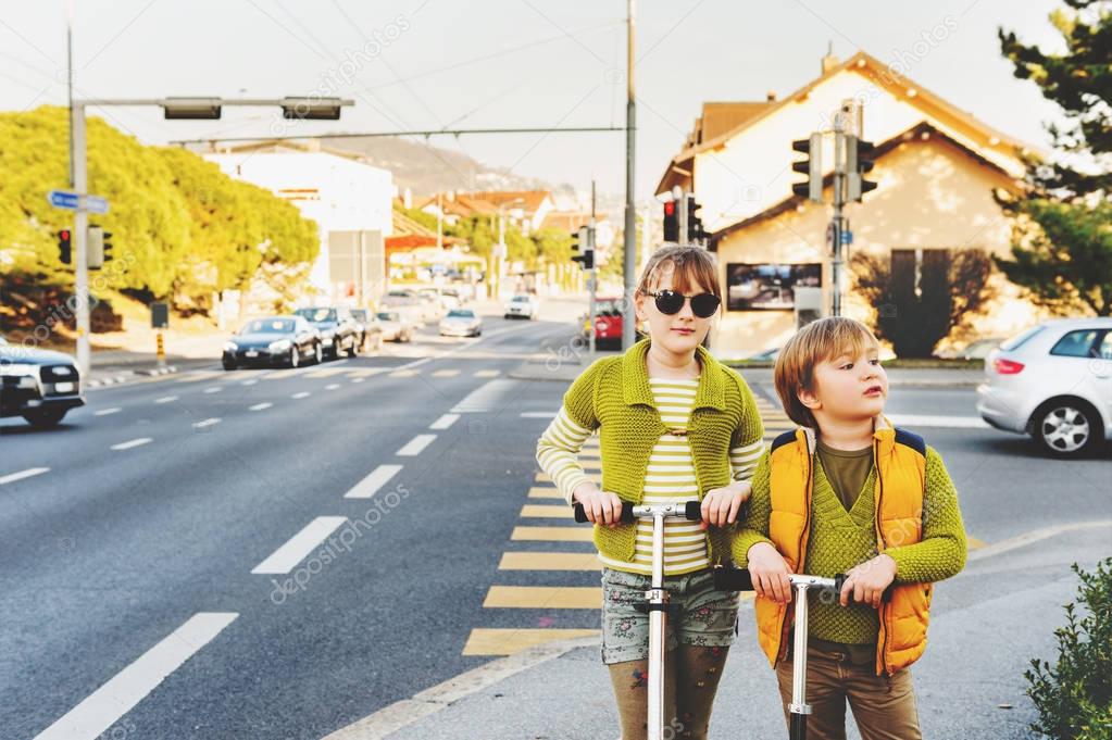 Two happy kids playing outdoors, fashion boy and girl posing on the street, riding scooters, crossing the road. Image taken in Lausanne, Switzerland