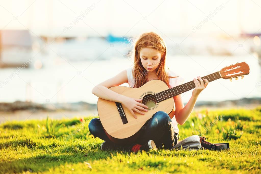 Outdoor portrait of adorable 9 year old kid girl playing guitar outdoors, sitting on bright green lawn at warm summer sunset