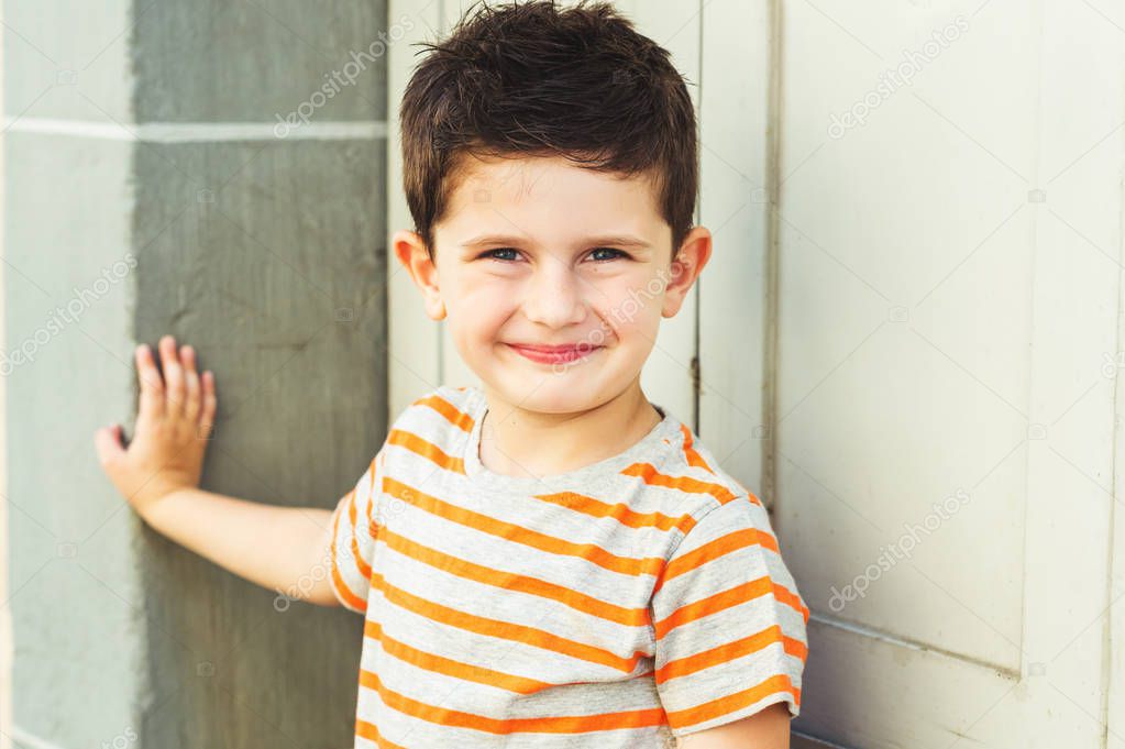 Outdoor portrait of a cute little boy standing against grey wall on a very sunny day, wearing grey and orange stripe tshirt