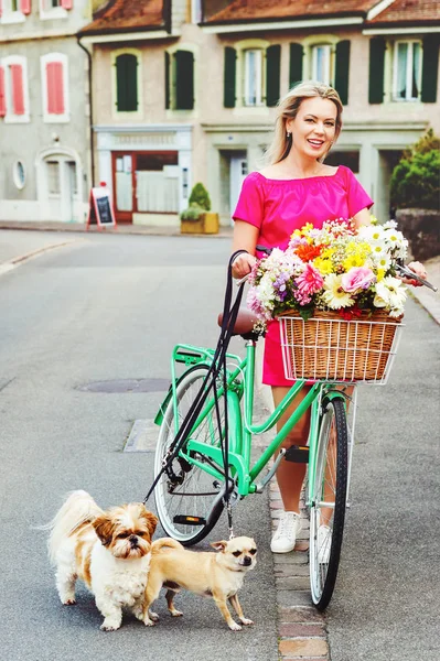 उज्ज्वल गुलाबी ड्रेस परिधान केलेली सुंदर गोरी स्त्री, टोपलीत मोठ्या रंगाच्या फुलांचा पुष्पगुच्छ असलेली सायकल चालविणे आणि दोन लहान कुत्र्यांसह लीश ठेवणे. लॉसॅन, स्वित्झर्लंड मध्ये घेतले प्रतिमा — स्टॉक फोटो, इमेज