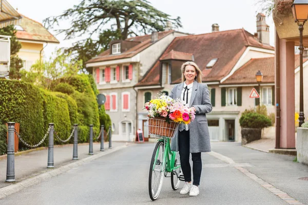 Hermosa mujer rubia con abrigo gris, montando una bicicleta con un gran ramo de flores de colores en la cesta. Imagen tomada en Lausanne, Suiza — Foto de Stock