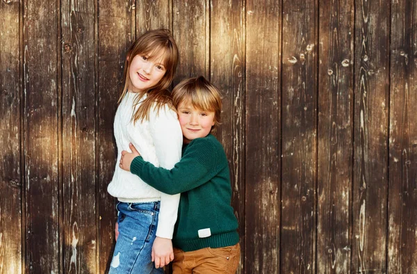 Outdoor Portret van twee schattige kinderen, kleine broer en grote zus, dragen van gebreide pullovers, permanent tegen houten achtergrond in zonlicht — Stockfoto