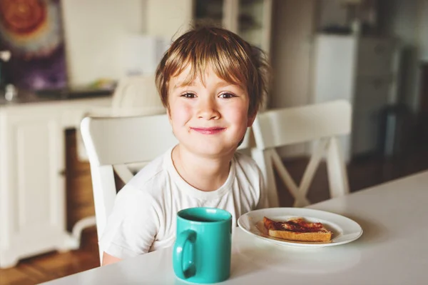 Bonito menino comendo sua torrada com geléia e chocolate quente para o café da manhã — Fotografia de Stock